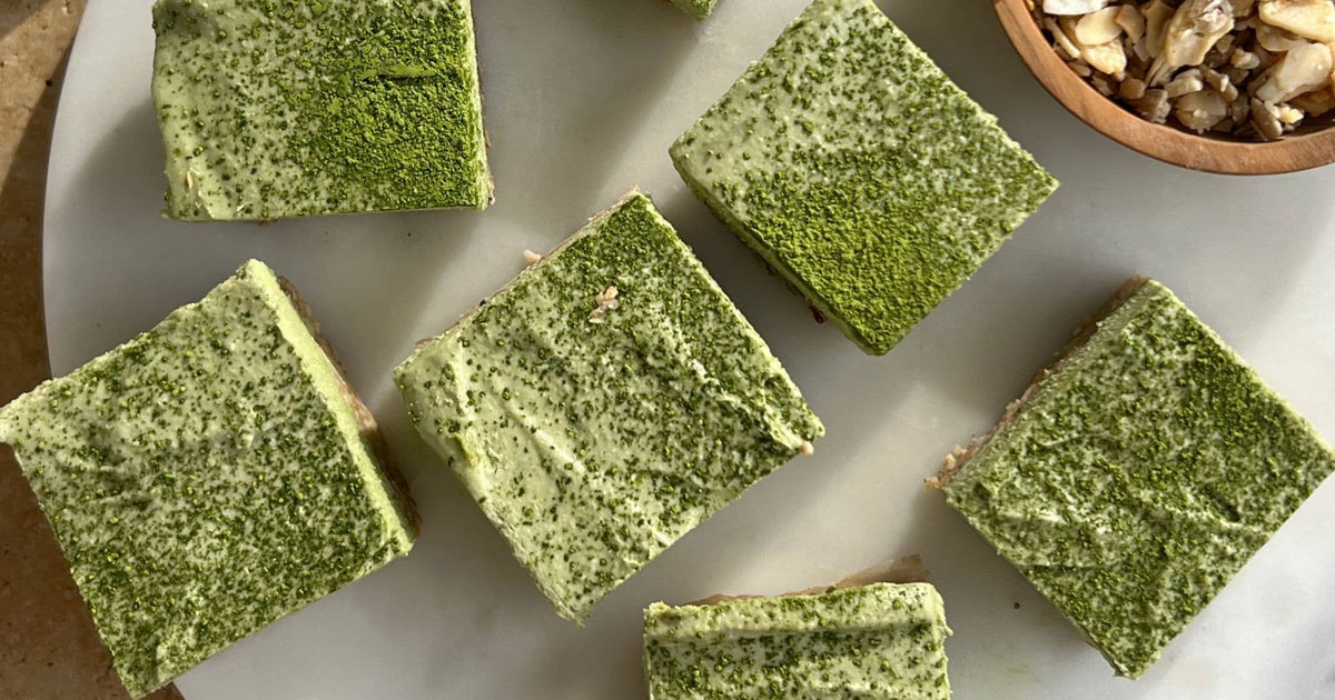 Sliced matcha peppermint cheesecake bars dusted with matcha powder on a marble board, with a bowl of nuts alongside