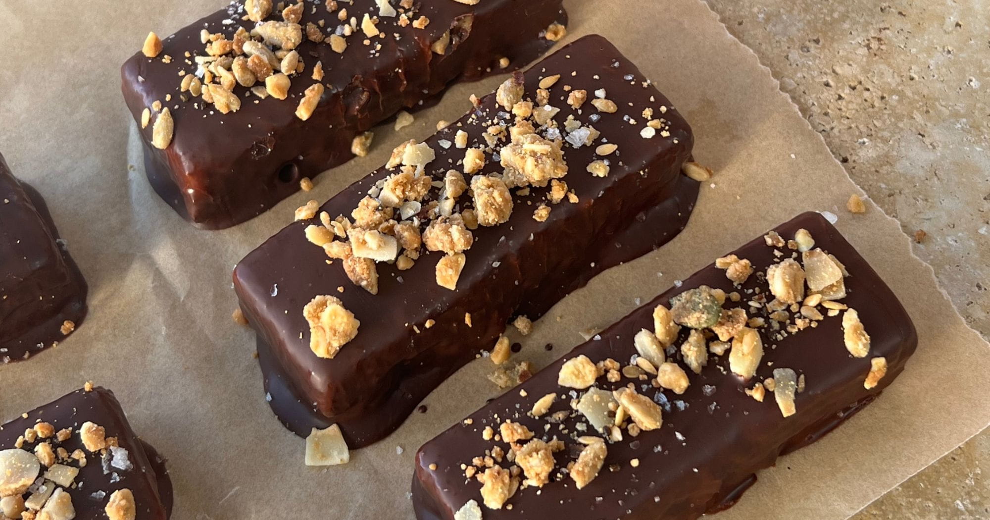 Grain-free protein peanut caramel bars coated in dark chocolate and topped with crushed peanuts and Paleo Foods Co Protein Boost granola, on baking paper