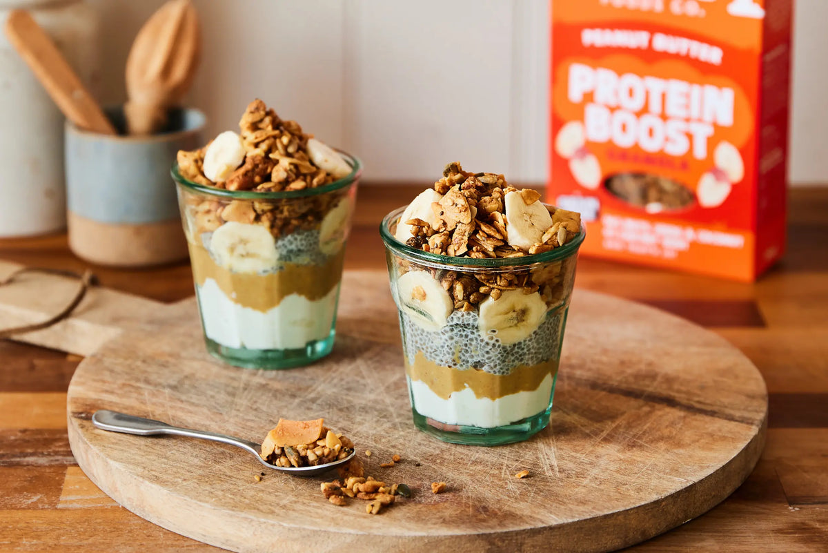 Two layered protein smoothie cups topped with banana slices and granola, served on a round wooden board with a spoon of Paleo granola, and a red box labeled Protein Boost in the background.