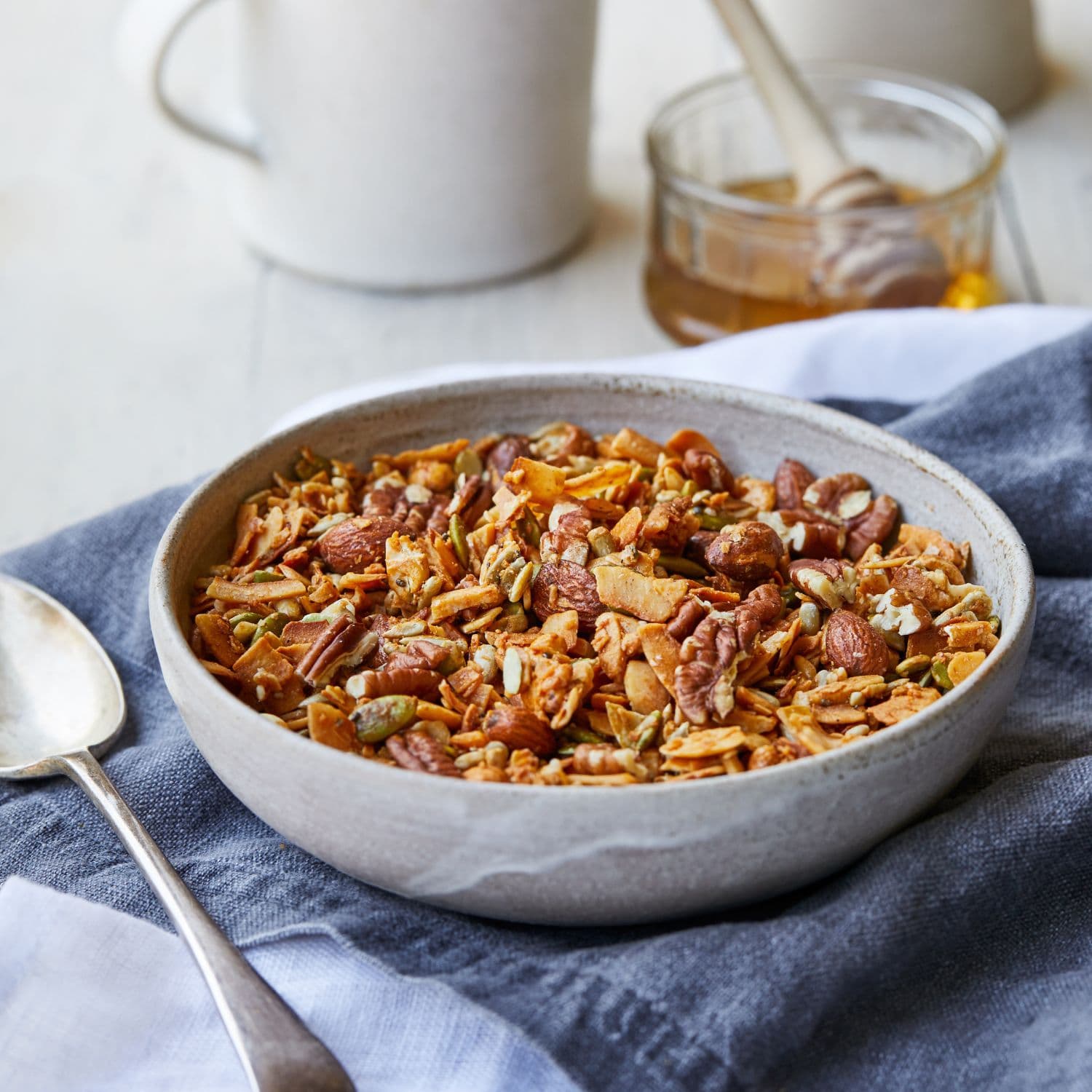 A bowl of Paleo granola with nuts, seeds, and dried fruit, set on a blue cloth with a spoon and a jar of honey in the background.