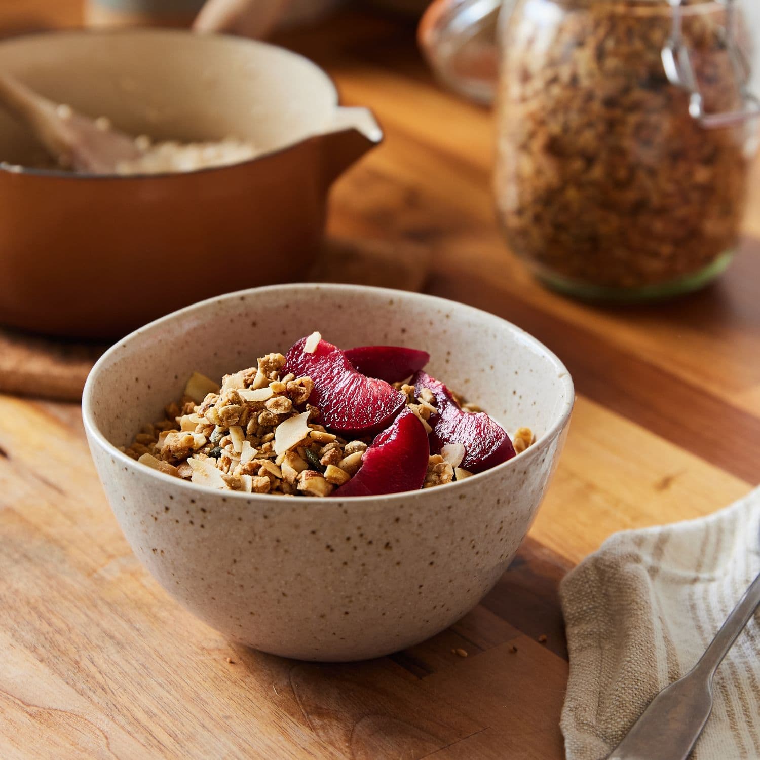 A speckled beige ceramic bowl filled with Paleo granola and dark red fruit slices, set on a wooden table with a blurred jar of granola and a spoon in the background.