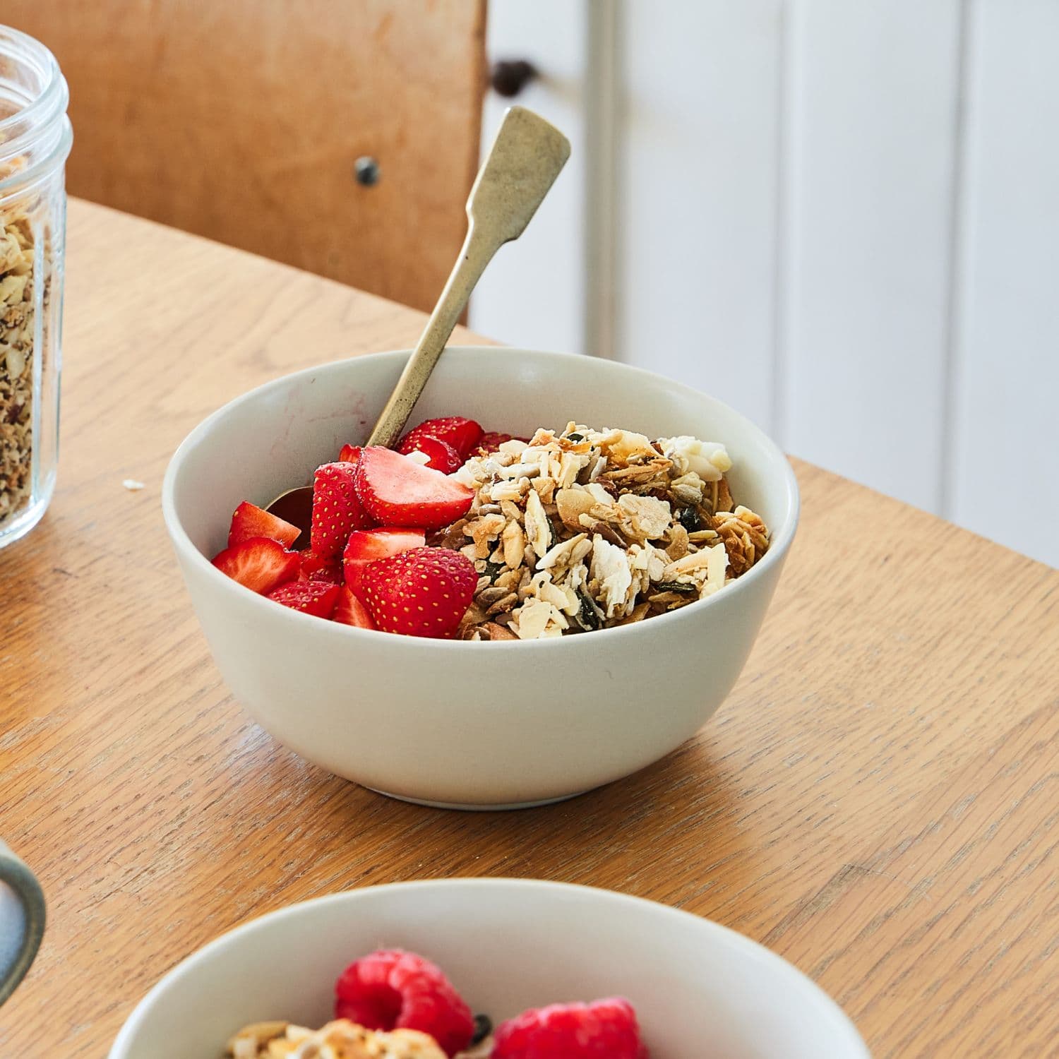 A light gray ceramic bowl filled with Paleo granola, sliced strawberries, and a wooden spoon, set on a wooden table in a bright indoor setting.