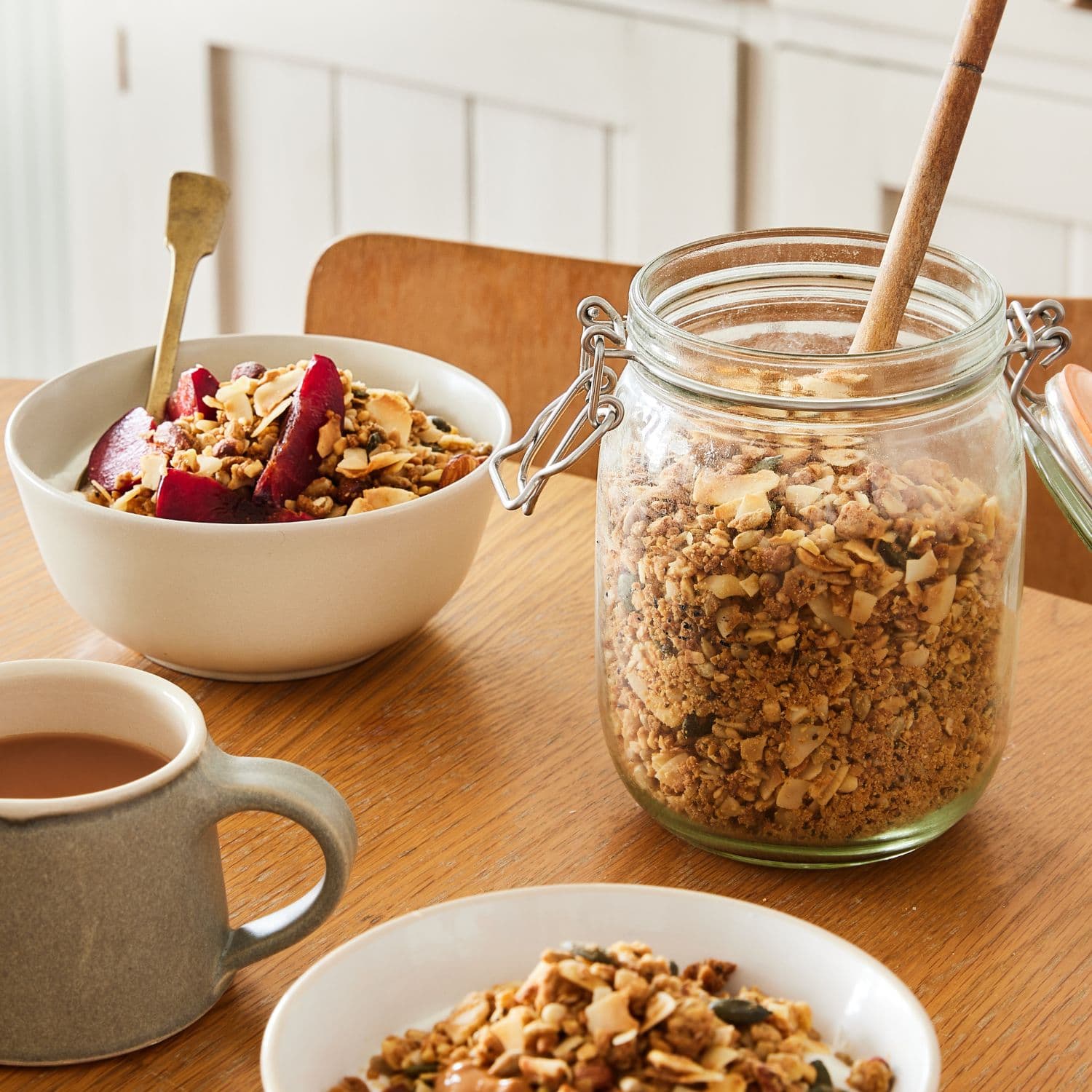 Paleo granola in a clear glass clamp-lid jar with a wooden spoon, shown beside bowls of granola and a mug on a wooden table.