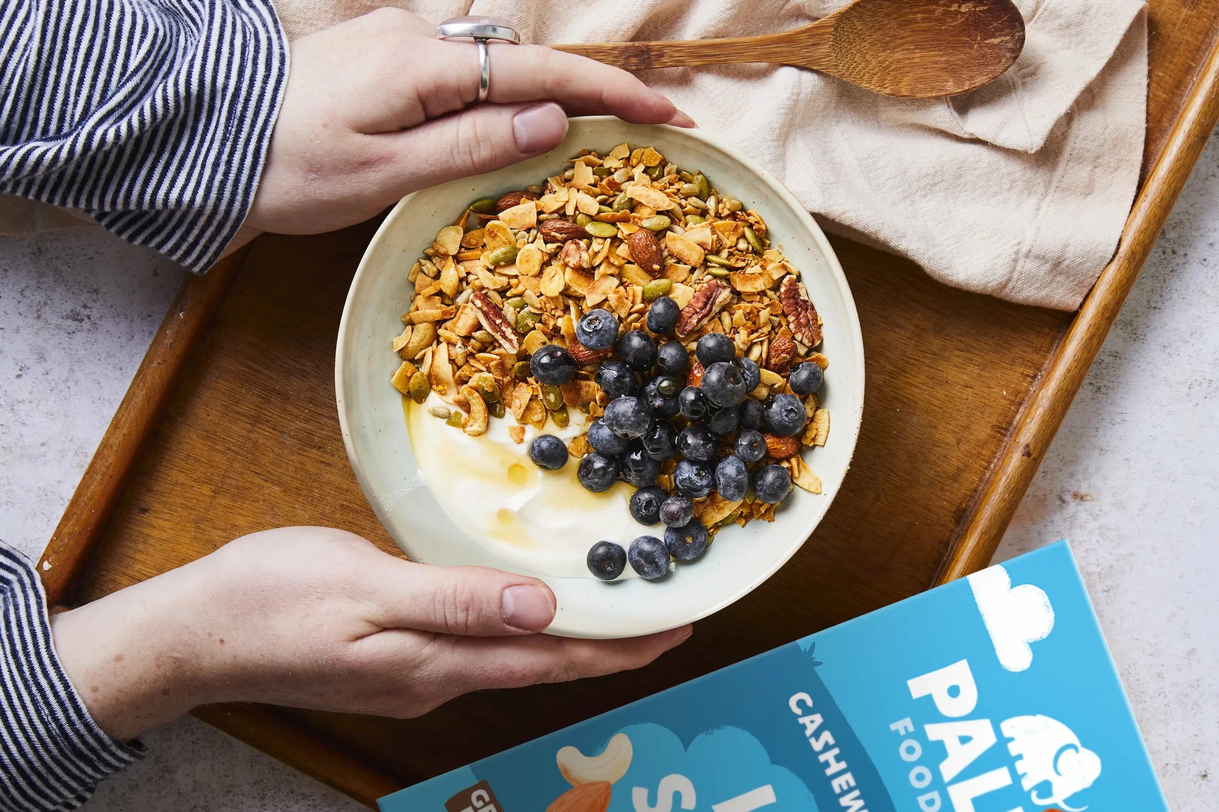A top-down view of a bowl of granola with yogurt and blueberries held by hands, placed on a wooden tray with a blue box of Palmetto Foods Cashew Flakes in the foreground.