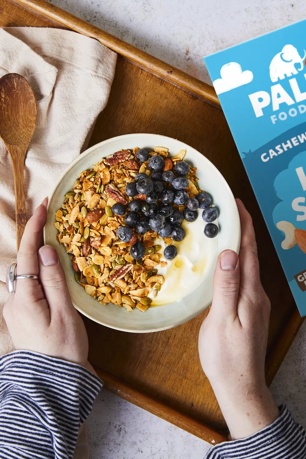 A top-down view of a bowl of granola with yogurt and blueberries held by two hands over a wooden tray, with a blue box of Paleo Foods Cashew Protein Granola visible on the right.
