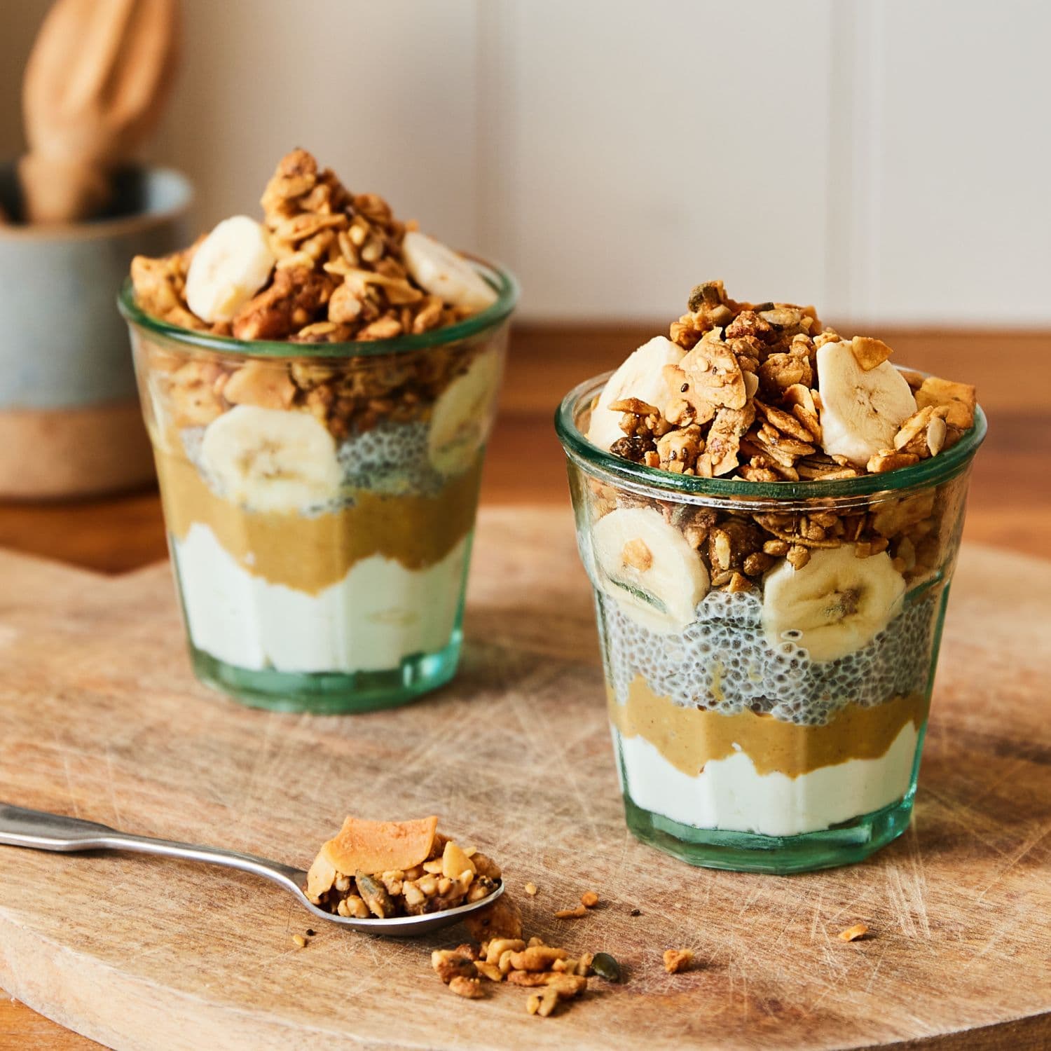 Two layered granola yogurt cups topped with banana slices and Paleo granola, arranged on a wooden board with a spoon in the foreground.