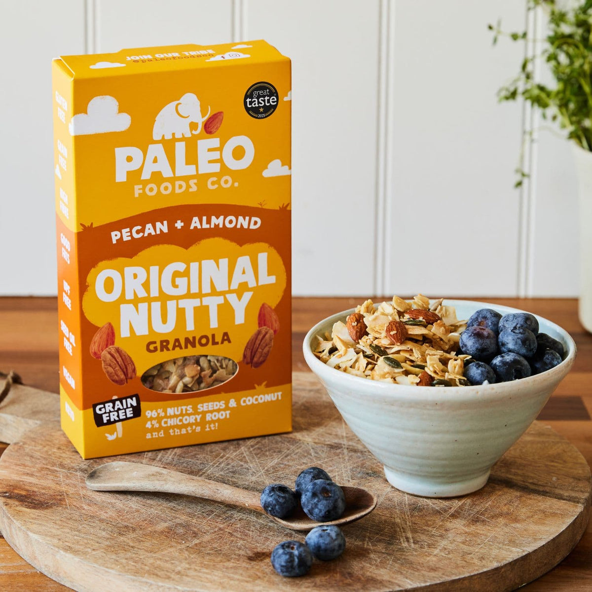 Box of Paleo Foods Co. pecan and almond granola with a bowl of granola and blueberries on a wooden board.